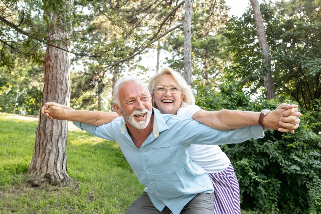 Happy senior couple smiling and playing together outdoors, with the man carrying the woman on his back in a park setting.